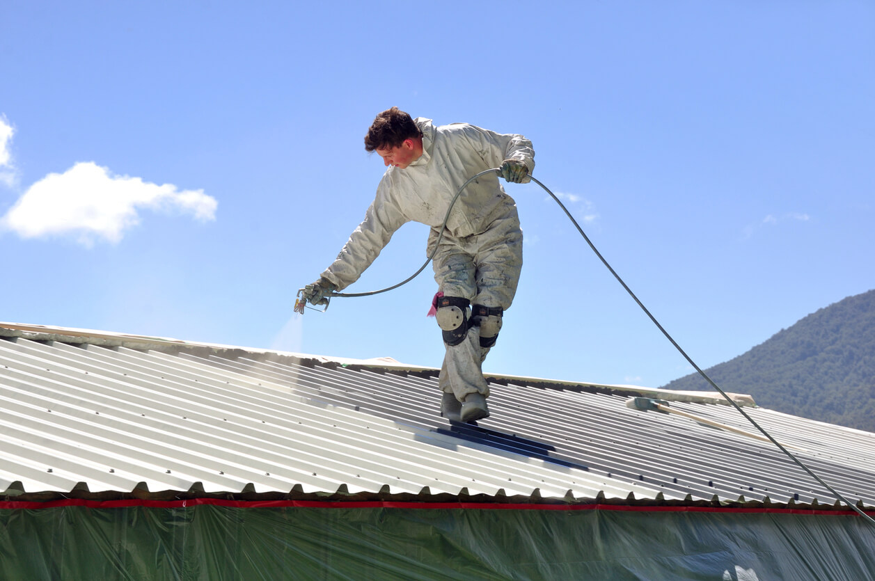 Home remodeling expert using a spray gun to apply white paint on a metal roof.