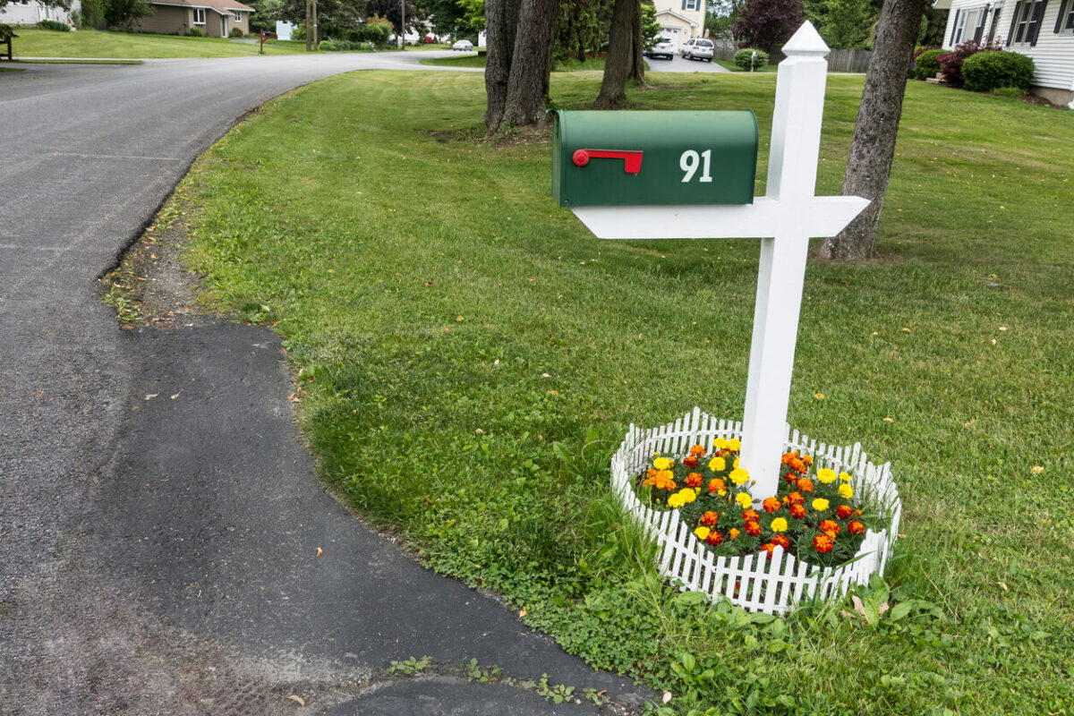 Green mailbox at the end of a driveway with a small garden of yellow and orange flowers surrounded by a small picket white fence at the bottom of the post